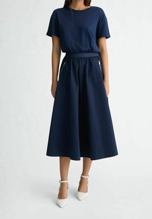 Mujer con una blusa de manga corta azul marino metida en una falda midi de pierna ancha y cintura alta, con bolsillos con cremallera en la parte delantera y tacones blancos con tira en el tobillo.