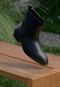 Schwarze Lederstiefelette mit niedrigem Absatz, die über einer Holzbank im Freien steht, mit Gras und Pflanzen im Hintergrund.