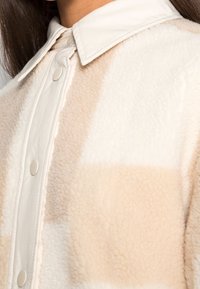 Primer plano de una chaqueta de sherpa a cuadros beige y blanca con un botón blanco y un suave cuello, llevada por una persona con cabello largo y oscuro.