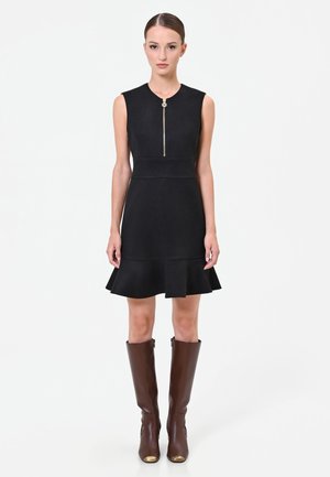 Donna in piedi che indossa un vestito nero senza maniche con orlo svasato e cerniera frontale, abbinato a stivali di pelle marrone al ginocchio.