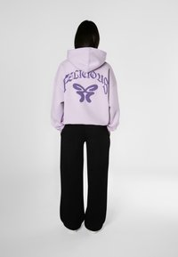 Levanduľová oversize mikina s nápisom "Fabulous" v tmavo fialovej farbe a grafikou motýľa na chrbte, spárovaná s čiernymi voľnými nohavicami.