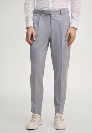 Mann trägt hellgraue, schmal geschnittene, karierte Hose, kombiniert mit weißen Turnschuhen und einem in die Hose gesteckten weißen Hemd, steht vor einem einfarbigen Hintergrund.