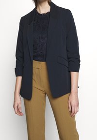 Námořnická blazer s hladkou texturou, se zářezovými klopy a vyhrnutými rukávy, nošená přes krajkový top, doplněná hnědými kalhotami s vysokým pasem.