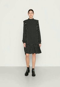 Vestido preto com bolinhas brancas, mangas longas e ombros franzidos, apresentando uma bainha em camadas. Usado com botins pretos. Textura de tecido suave.