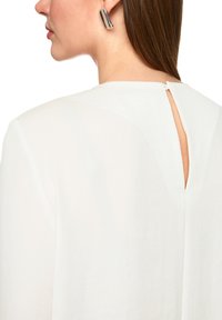 Weißes Top mit Ausschnitt am Rücken, rundem Ausschnitt und weichem Stoff. Das Model trägt silberne geometrische Ohrringe.