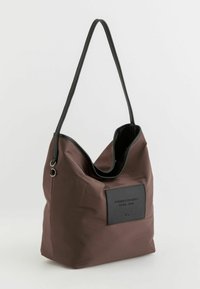 Brun nylon skuldertaske med en fleksibel form, sort syntetisk kant og en rektangulær sort lap med præget tekst på forsiden.