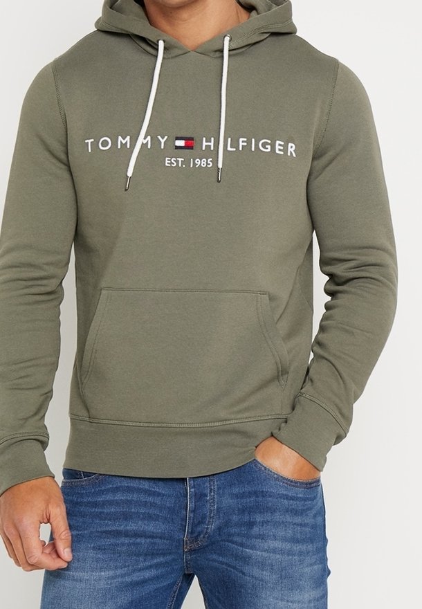 Homme portant un sweat à capuche Tommy Hilfiger vert olive avec cordons blancs et poche avant, associé à un jean bleu, main dans la poche.