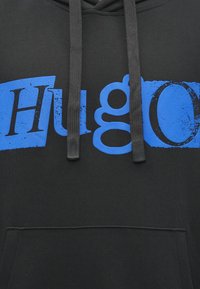 Felpa nera con tasca frontale e lettere blu a blocchi che mostrano parzialmente la parola "Hugo" in uno stile consumato, due lacci neri visibili.