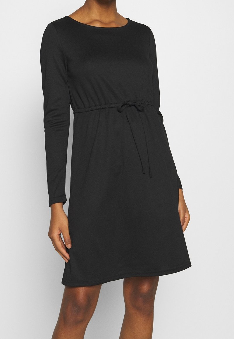 Femme portant une robe noire mi-longue à manches longues avec un col rond et un détail de ceinture nouée, sur un fond blanc uni.
