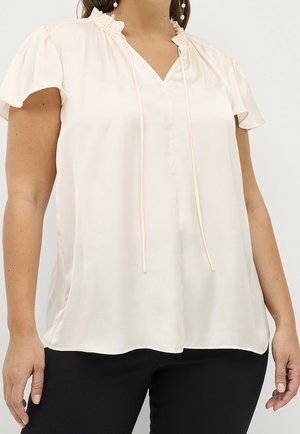 Kvinde iført en cremefarvet, kortærmet bluse med flæsekant ved halsen og snører, kombineret med sorte bukser.