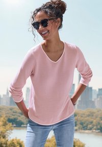 Light pink knit long-sleeve shirt with a v-neck and rounded hem, paired with blue denim jeans. Features curly hair and dark sunglasses.