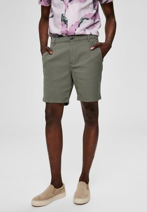 Homme portant un short vert olive, une chemise rose à manches courtes à motifs rentrée, et des chaussures beige sans lacets, debout les mains dans les poches.