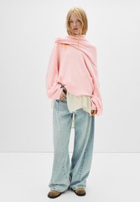 Svetlo roza oversized pulover z dolgimi rokavi, oblečen preko bele teksturirane majice, v kombinaciji z širokimi modrimi kavbojkami in rjavimi natikači.