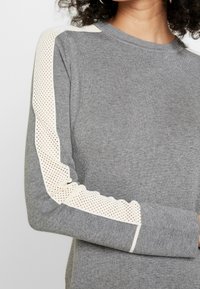 Maglione grigio con maniche lunghe, caratterizzato da un tessuto testurizzato e un dettaglio in rete beige lungo le maniche esterne per la ventilazione.