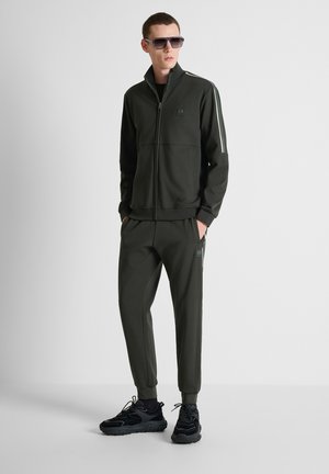 Uomo che indossa una giacca sportiva nera con zip e pantaloni coordinati con scarpe sportive e occhiali da sole, in piedi davanti a uno sfondo semplice.