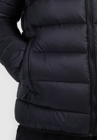 Personne portant une doudoune noire avec une main dans la poche latérale, matelassage horizontal visible et fermeture zippée sur le devant.