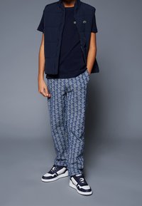 Marinblå pufferväst över en marinblå t-shirt, mönstrade blå byxor och vita sneakers med marinblå detaljer. Byxorna har en geometrisk design.