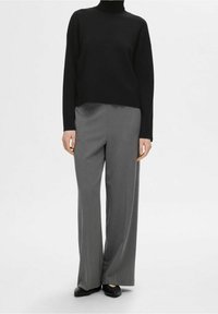 Zwarte coltrui met lange mouwen gecombineerd met lichtgrijze, wijde pantalons en zwarte platte schoenen, op een effen witte achtergrond.