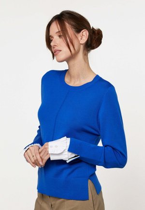 Femme aux cheveux bruns attachés en chignon, portant un pull bleu vif par-dessus une chemise blanche avec des poignets à volants, pantalon beige, regardant vers le bas, mains croisées.