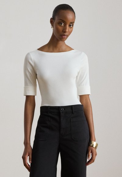 Femme à la peau foncée et aux cheveux attachés porte un haut blanc à encolure bateau avec manches retroussées et un pantalon noir taille haute sur fond uni.