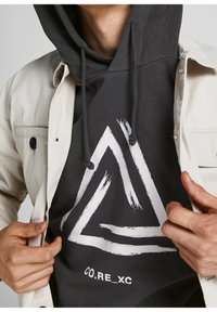 Sudadera con capucha de color gris oscuro con un gráfico triangular abstracto en blanco, con cordones, llevada debajo de una chaqueta de color claro. El texto dice "CO.RE_XC."