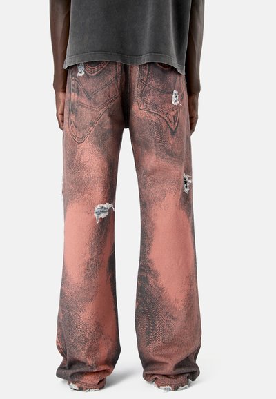 Loszittende roze en zwarte distressed jeans met scheuren op zakken, benen en rafelige zomen, gedragen door een persoon die met de rug naar de camera staat.