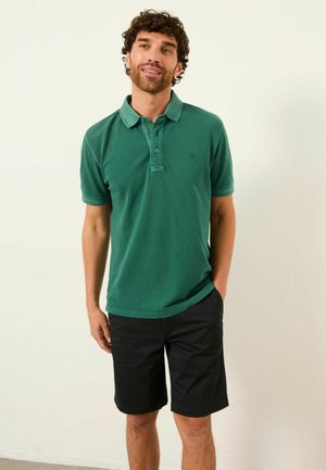 Homme aux cheveux bouclés portant un polo vert à manches courtes et un short noir, debout avec une main dans la poche.