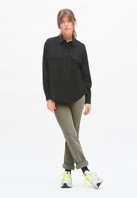 Black button-up shirt with two chest pockets, paired with cuffed olive green pants and gray sneakers with neon accents. Plain white background.