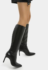 Svarta läder knähöga stövlar med spetsig tå och stilettklack. Har snörningsdetalj längs sidan och en slät, glänsande yta.