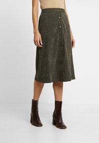 Oliven grøn nederdel i blødt stof, A-linjeformet, med en frontlukning med mørkebrune knapper. Stylet med brune hælede støvler.