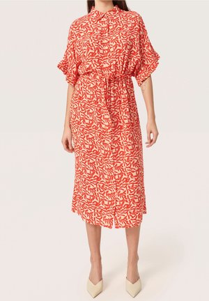 Woman wearing a buttoned, collared midi dress with orange leaf pattern, puffed sleeves, and beige pointed-toe heels against plain background.