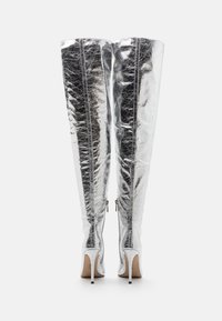 Botas plateadas por encima de la rodilla con un acabado metálico brillante. Presentan un tacón de aguja, cremallera lateral y una textura suave. Costura detallada en la parte posterior.