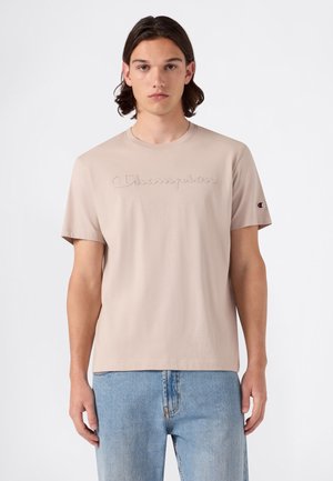 Młody mężczyzna z brązowymi włosami do ramion, ubrany w beżowy t-shirt Champion i jasnoniebieskie jeansy, stoi na tle jednolitego białego tła.