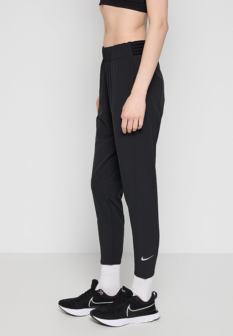 Pantalons de sport noirs avec une taille texturée, des jambes effilées et un logo Nike réfléchissant près de la cheville, associés à des baskets noires et des chaussettes blanches.