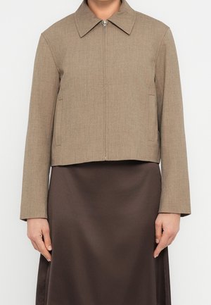 Kvinde iført en lysebrun jakke med lynlås og krave samt en mørkebrun satin nederdel, vist fra hals til midt på låret.