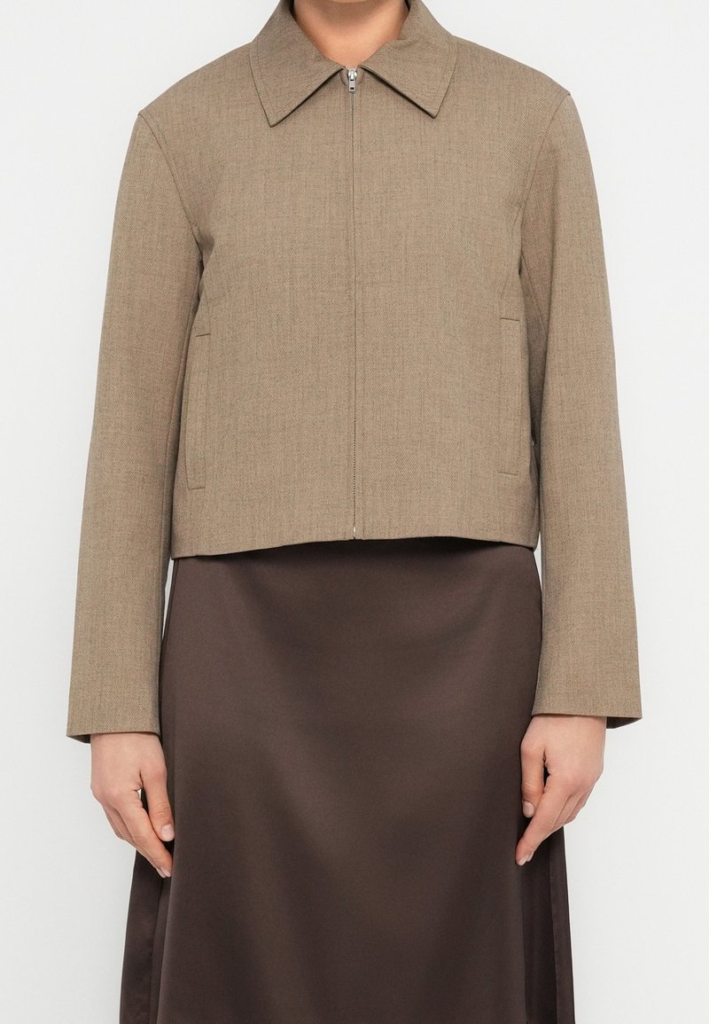 Femme portant une veste zippée marron clair avec col et une jupe en satin marron foncé, montrée du cou jusqu’au milieu de la cuisse.