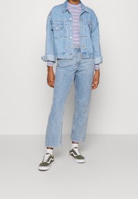 Světle modrá džínová bunda s volným střihem a ohrnutými rukávy, v kombinaci se světle modrými džíny. Bílá a olivově zelená teniska doplňuje celý outfit.