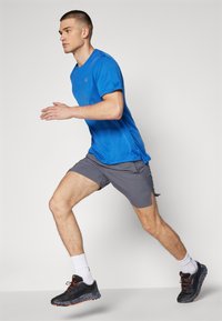 Camiseta deportiva azul, pantalones cortos grises, zapatillas de correr negras con detalles naranjas. Modelo en movimiento, mostrando un diseño atlético y en forma.