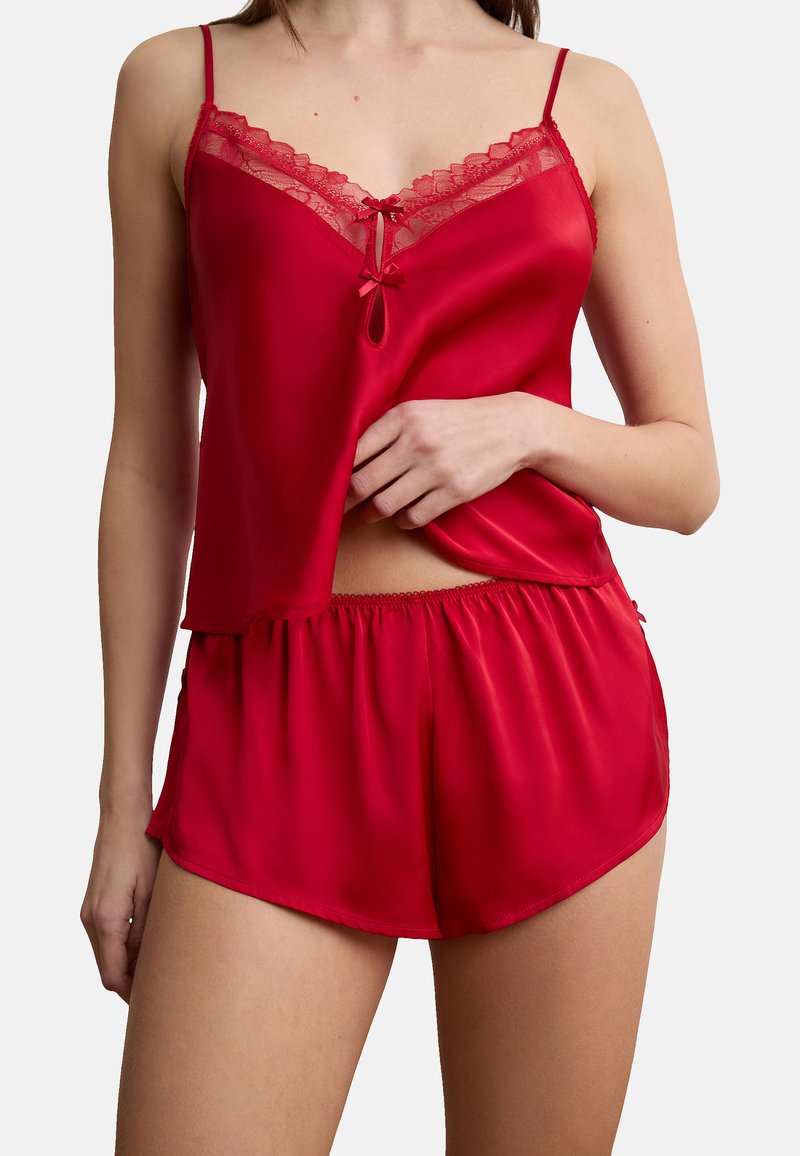 Frau trägt rotes Satinsamt-Camisole mit Spitzenbesatz und passende Shorts, Hand hebt den Saum des Camisole leicht an, vor weißem Hintergrund.