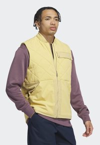 Prešívaná žltá vesta so zipsom na prednej strane, má náprsné vrecko a pod ňou je kontrastná fialová košeľa s dlhým rukávom. Nosená spolu s námorníckymi nohavicami.