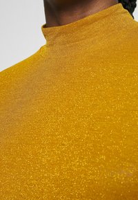 Goldener, funkelnder Rollkragenpullover mit hohem Kragen, aus strukturiertem Stoff. Verfügt über einen Schimmereffekt mit feinem Glitzer im gesamten Material.