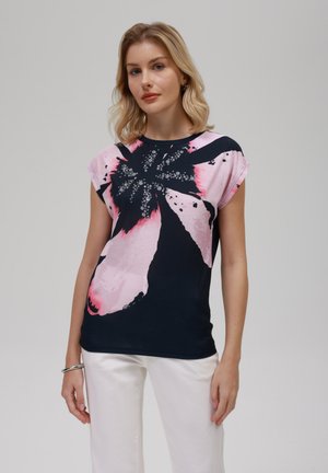 Lyshåret kvinde iført en sort og pink abstrakt blomstret ærmeløs top og hvide bukser, stående mod en ensartet baggrund.