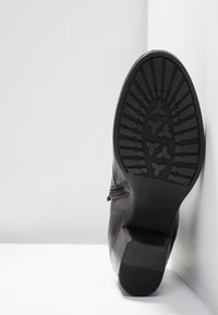 Semelle d'une botte en cuir noir avec motif sur la semelle, talon bloc et fermeture éclair latérale, positionnée debout contre un mur d'angle blanc.