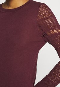 Nahaufnahme eines burgunderfarbenen Pullovers mit Rundhalsausschnitt und halbdurchsichtigen Ärmeln im Spitzenmuster.
