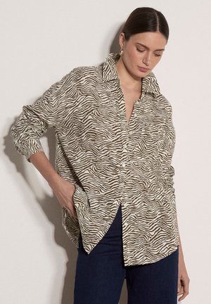 Frau trägt ein übergroßes, beiges und braunes Hemd mit Zebra-Muster und Knöpfen, dazu dunkle Hosen, steht vor einem schlichten hellen Hintergrund.