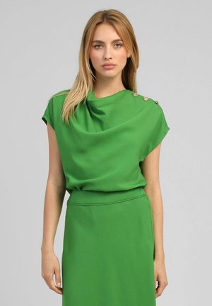 Femme blonde portant un haut sans manches vert avec des détails boutons sur l'épaule et une jupe verte assortie sur un fond gris.