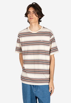 Young man with medium-length hair wearing a cream T-shirt with brown, black, and red horizontal stripes, and blue jeans against a plain background.