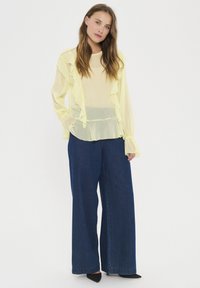 Durchsichtige gelbe Bluse mit fransigen Kanten, kombiniert mit weit geschnittenen, dunkelblauen Jeans. Schwarze spitze Absätze runden das Outfit ab.