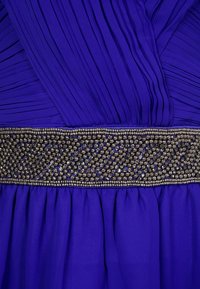 Fialové šaty s plisovanou tkaninou, zdobeným pasem s jemnými korálky a splývavou strukturou.