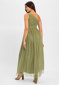 One-shoulder olive green dress with ruched bodice and flowing tulle skirt. Paired with green heeled sandals. Model shown from back.